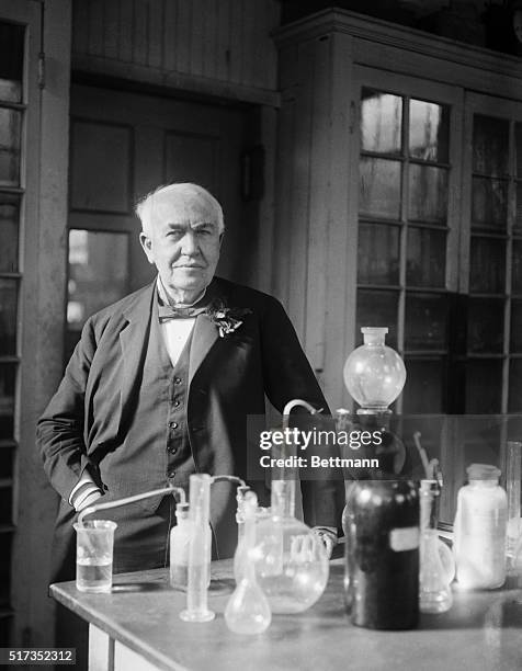 Thomas Edison standing in his lab. Edison is standing behind a table that has various beakers and equipment. His hand is resting on the table....