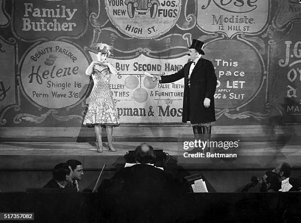 Vaudeville performers on stage. On the right the woman appears dejected while the man in top hat, overcoat and boots, tries to hand her a rolled...