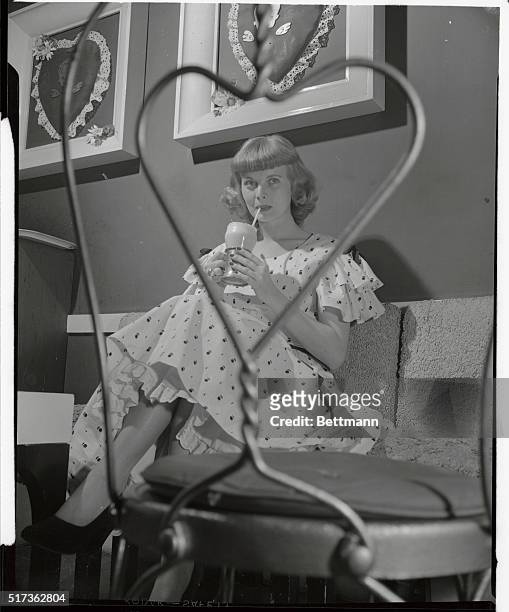 Looking demure as an old-fashioned valentine, Dorothy Abbott, drinking an ice-cream soda, is seen through the frame of a wire-back chair. Her pink...