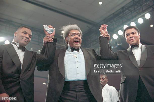 Mike Tyson, Don King, and Mohammed Ali, all clasping hands wearing tuxedos in the boxing ring at the Las Vegas Hilton.
