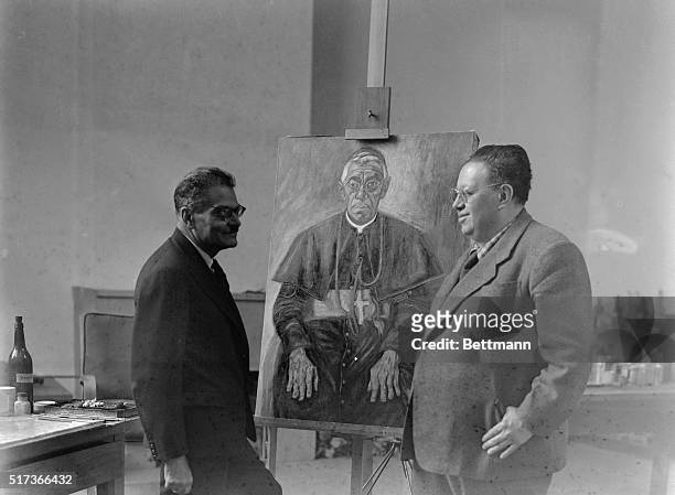 Photo shows the Mexican painter Diego Rivera, , standing with muralist Jose Clemente Orozco, , in front of a portrait of an Archbishop.