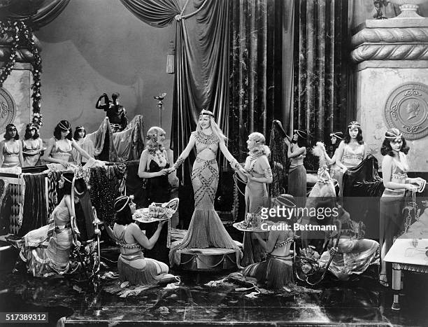 Publicity handout of a movie still of Claudette Colbert in the 1934 movie, "Cleopatra." Colbert stands on a pedestal in the middle of a large, lush....