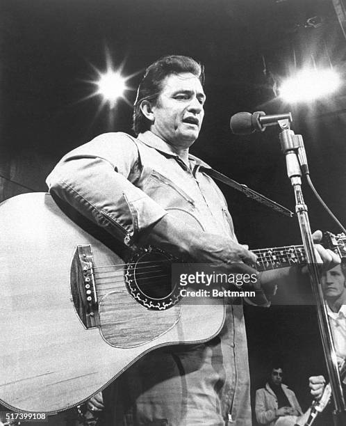Picture shows country singer, Johnny Cash, performing on stage. He is playing the guitar and singing into a microphone.