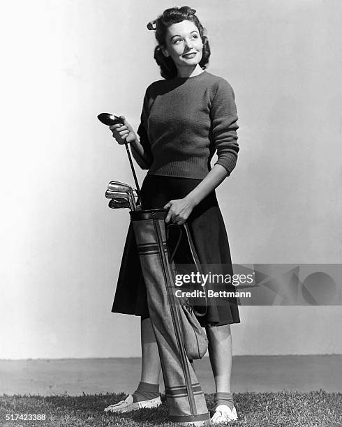 Woman holding a golf club and a golf bag, and a golf ball is lying on the ground. Full length photograph. Ca. 1940s-1950s.