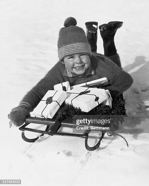 Young boy lays on a sled laden with Christmas presents and evergreen wreath. Undated photograph circa 1950.