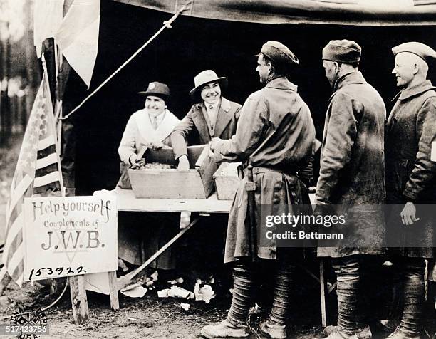 Women at a World War I American embarkation center, in France, handing out chocolates, cookies, and cigarettes to US soldiers near a sign that reads,...