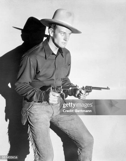 Actor John Wayne is shown in a publicity handout from the 1930s or 1940s, holding two guns and wearing typical western gunslinger garb. 3/4 length...