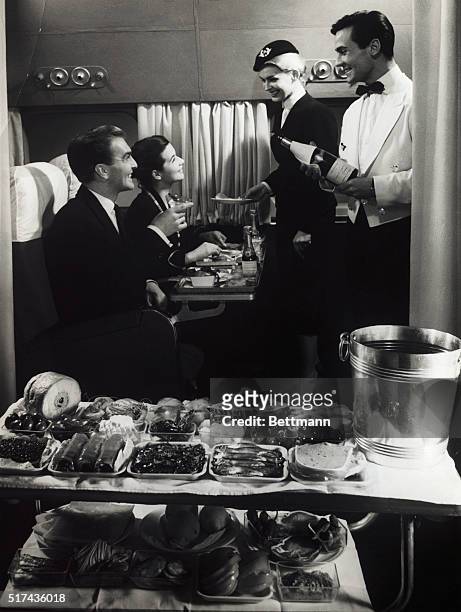 Undated photograph of a couple being served luxoriously by a steward and stewardess aboard an airplane.