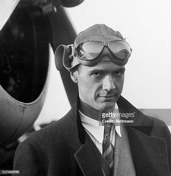 Howard Hughes standing in front of an airplane in a leather flight helmet and goggles.
