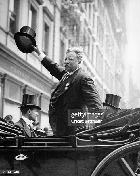 President Theodore Roosevelt waves his hat while standing in an automobile.