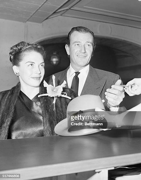 Hollywood, CA-: An attractive hat-check girl at Club Mocambo in Hollywood holds the attention of Mr. And Mrs. John Wayne. Mrs. Wayne is the former...