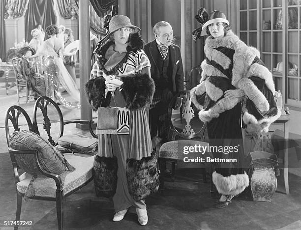 Two women, visiting a dress salon, donning styles of the late 1900s, including broad-brimmed, ribboned hats, fur trimmed cloaks over ankle-length,...