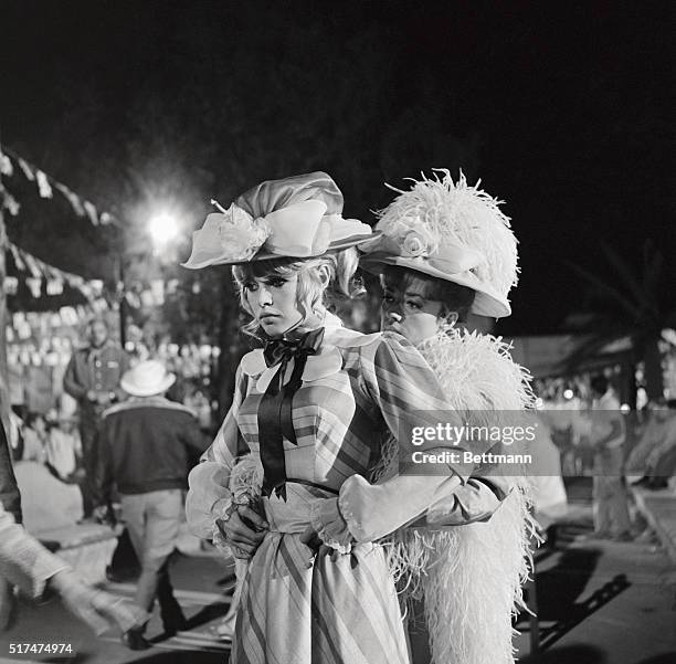 Dressed in the regalia of turn-of-the-century costumes, Brigitte Bardot receives a helping hand from her co-star, Jeanne Moreau, on the set of Viva...