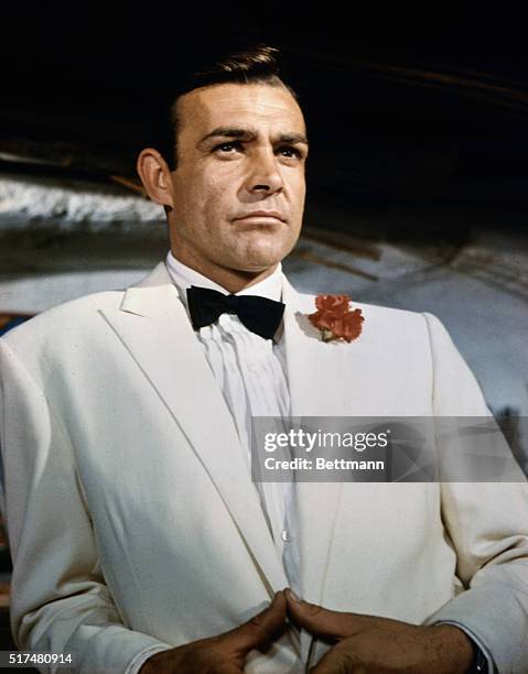 Ca. 1966: Waist-up portrait of Sean Connery, as James Bond, leaning against a bar and looking out across the room. Connery is wearing a white tuxedo...