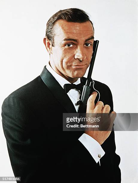 Waist-up portrait of Sean Connery, as James Bond, caressing the barrel of a gun against the side of his face. Connery is wearing a tuxedo and bow tie...
