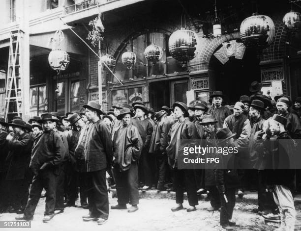 Group of men in Asian jackets and shoes wear hats and stand on the sidewalk and in the street below paper lanterns outside the Yuen Fat & Co....