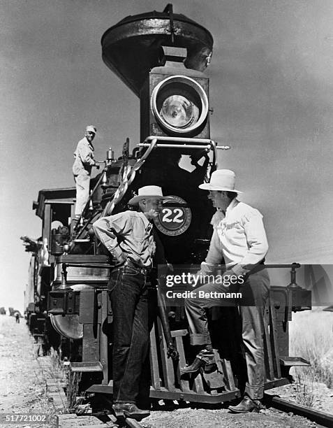 Director Howard Hawks and John Wayne chat between scenes while standing infront of old-fashioned train.