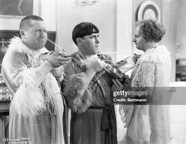 The Three Stooges comedy team, dressed in lady's dressing gowns, spray each other with seltzer water during a scene from one of their short films.