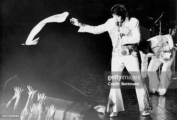 Elvis Presley, in a white jumpsuit with gold brocade and studs, tosses a nylon scarf into the waiting hands of the fans at a concert in the...