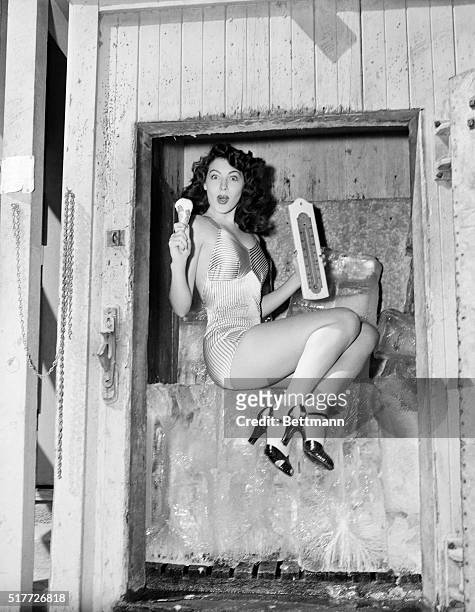 Actress Ava Gardner sits on a large block of ice while holding a thermometer in one hand and ice cream cone in the other.