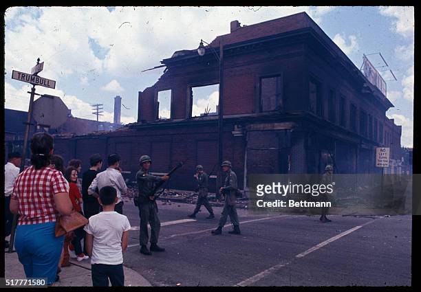Here is seen National Guard Troopers as they patrol a street corner at site of brick building, which is gutted and smouldering. Bystanders are...