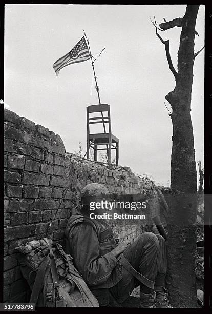 Marine radioman takes a breather along the wall of the Citadel in Hue. A makeshift flagpole and a chair support the American flag which flies over...