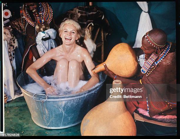 Actress Carroll Baker goes native and enjoys it as a Masai tribeswoman adds more water to her tin tub. The American actress used the facilities in a...