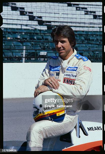 Rick Mears is shown sitting on his Indy car, wearing his racing outfit and holding his helmet in his lap.