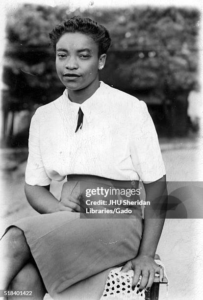 Portrait of an African American woman sitting on a bench outside with trees in the background, she is wearing a knee-length light colored skirt and a...