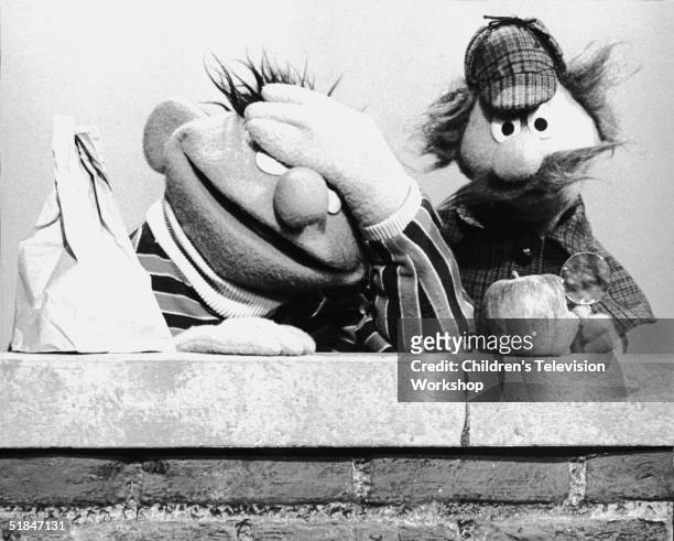 Muppet 'Ernie' holds his hand over his eyes in exasperation as 'Sherlock Hemlock' uses a magnifying glass to examine a bite taken out of an apple in...
