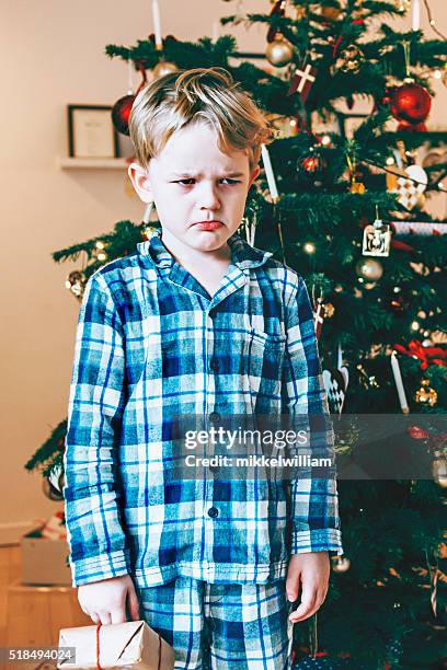 unhappy child in pajamas stands before christmas tree with present - christmas angry stock pictures, royalty-free photos & images