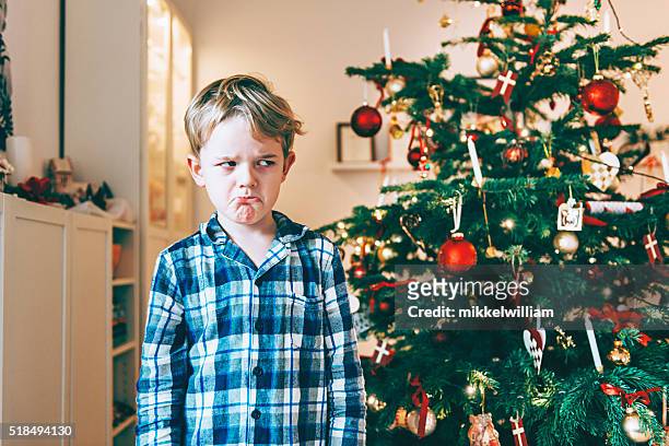 unhappy boy stands before chistmas tree and makes a face - sulking stockfoto's en -beelden