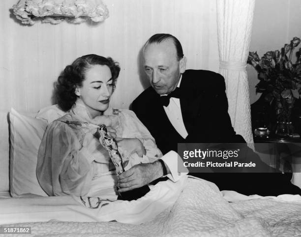 Man in a tuxedo presents the Best Actress Academy Award to American actress Joan Crawford for her performance in 'Mildred Pierce' as she lies in bed...