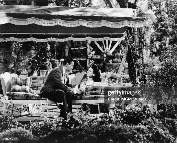 American actress Joan Crawford reclines and talks to English actor Herbert Marshall as he sits next to her on a bench swing before a water wheel in...