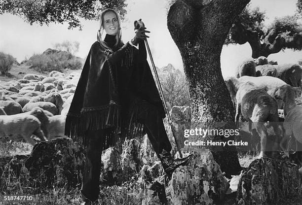 Maite de Soto Domecq stands under a tree, surrounded by sheep wearing a leather cape and matching pants by Loewe, near Extremadura, Spain.