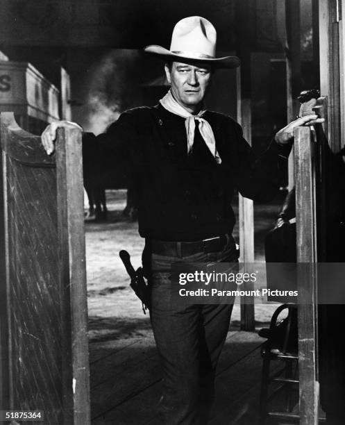 American actor John Wayne wears a Western outfit which includes a pistol in a holster and stands in a doorway in a still from the Cowboy film 'The...