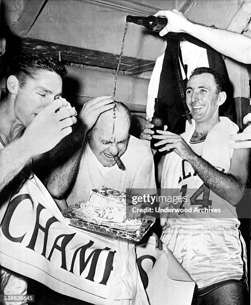 Bottle of beer is poured over Boston Celtic's coach Red Auerbach as Bill Sharman bites into a piece of victory cake and Bob Cousy smiles happily...