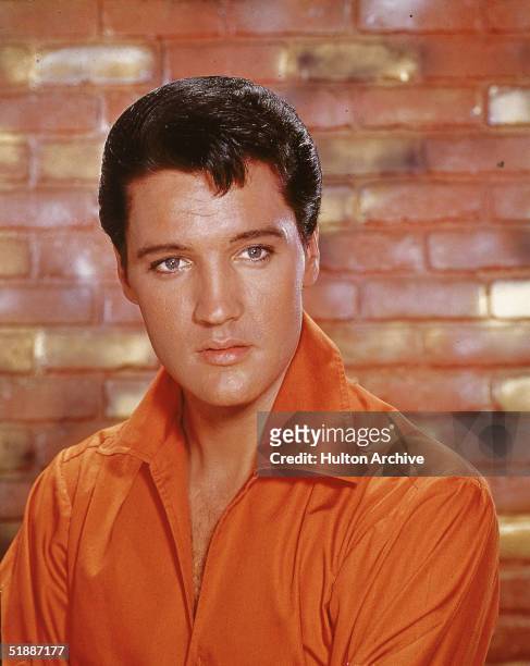 Portrait of American rock and roll singer Elvis Presley, dressed in an orange open-neck shirt and in front of a brick wall, mid 1960s.