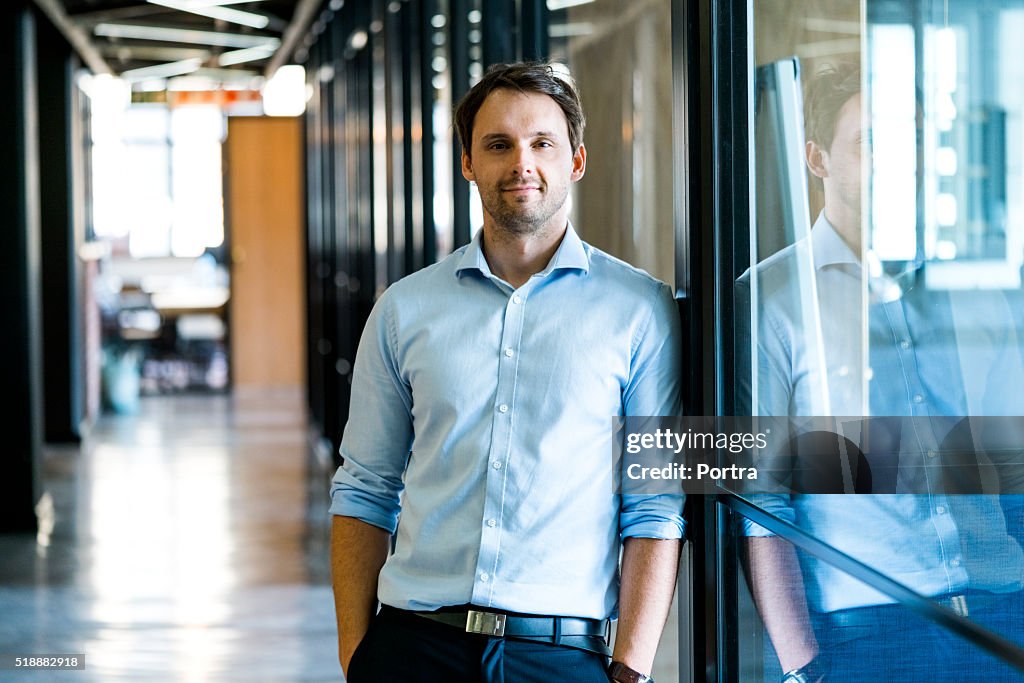 Confident businessman leaning on glass wall in office
