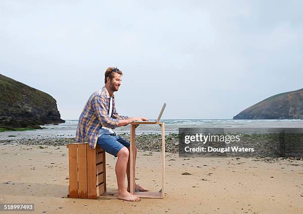 man using laptop on wooden crate at beach. - shorts stockfoto's en -beelden