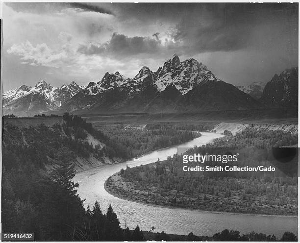 Black and white photograph of several of the peaks of the Teton Range covered in snow, the Snake River winds through the landscape and is surrounded...