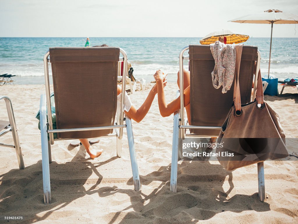 Two women sitting on beach chairs holding hands