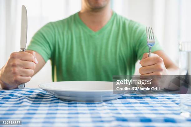 mixed race man holding fork and knife at table - hungry stock-fotos und bilder