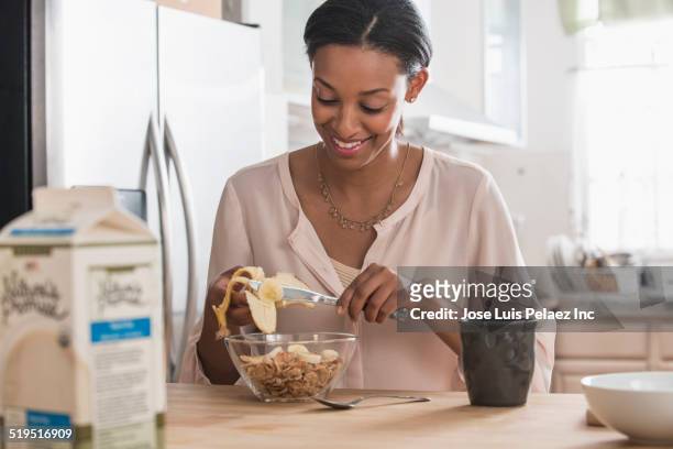 mixed race woman eating cereal and banana in kitchen - breakfast cereal fotografías e imágenes de stock
