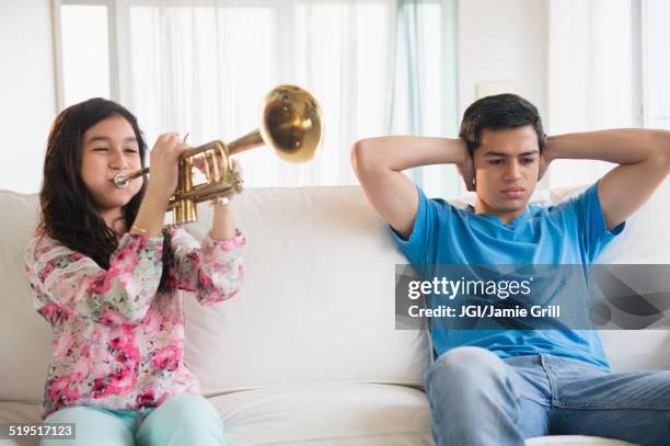 hispanic brother covering his ears as sister practices trumpet in living room - annoying brother stock pictures, royalty-free photos & images