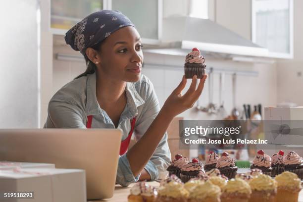 mixed race baker admiring cupcake in commercial kitchen - cupcake box stock pictures, royalty-free photos & images