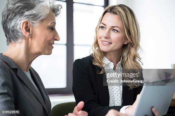 mid adult woman with tablet smiling at mature colleague - role model stock pictures, royalty-free photos & images