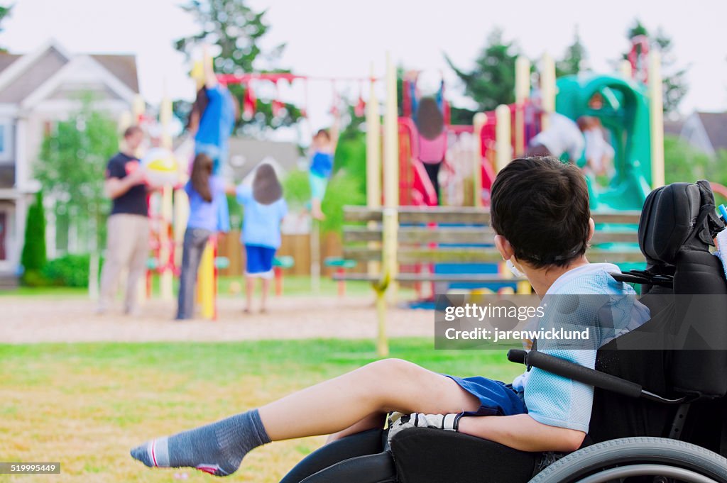 Disabled little boy in wheelchair watching children play on playground