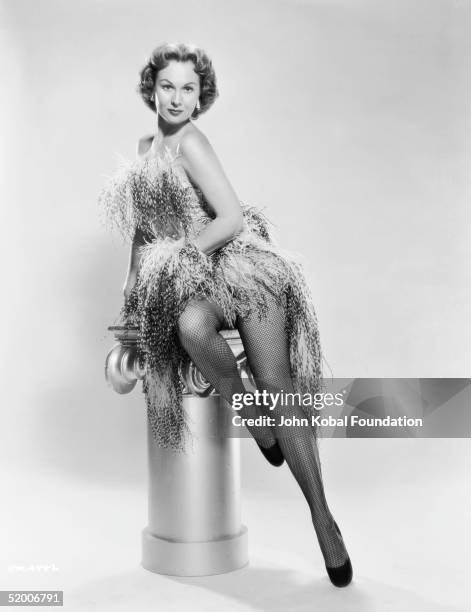 American film actress Virginia Mayo poses on a plinth, dressed in a feathery dress and fishnet stockings, circa 1950.