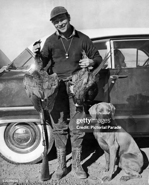 American actor and singer Roy Rogers leans against his Dodge car as he poses with two dead pheasants, his gun, and his hunting dog, early 1950s....